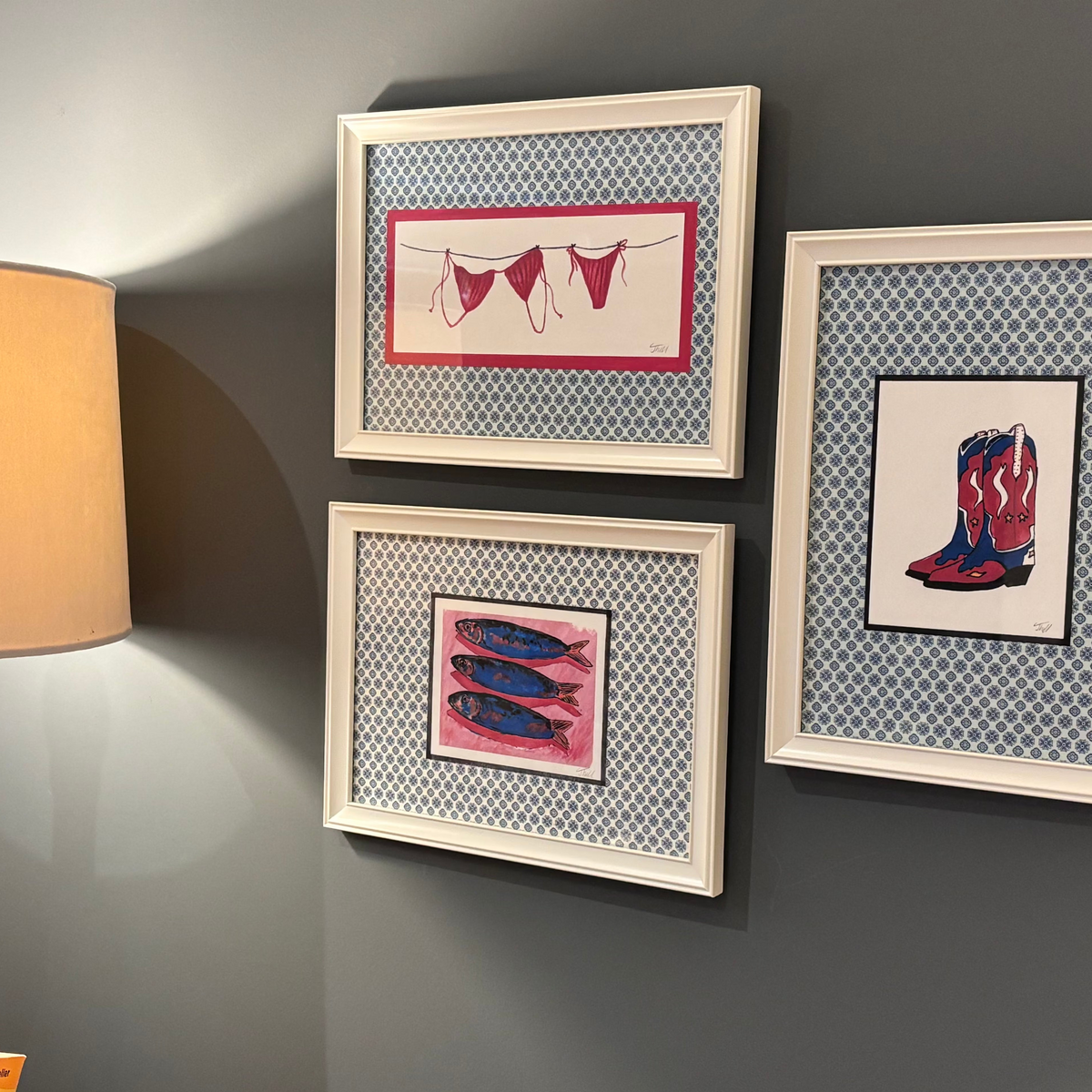 Three framed art prints displayed on a dark gray wall next to a lit lamp. The prints feature whimsical illustrations: underwear on a clothesline, three mackerel, and  a pair of cowboy boots, all set within a blue-and-white geometric patterned fabric and white wooden frames.