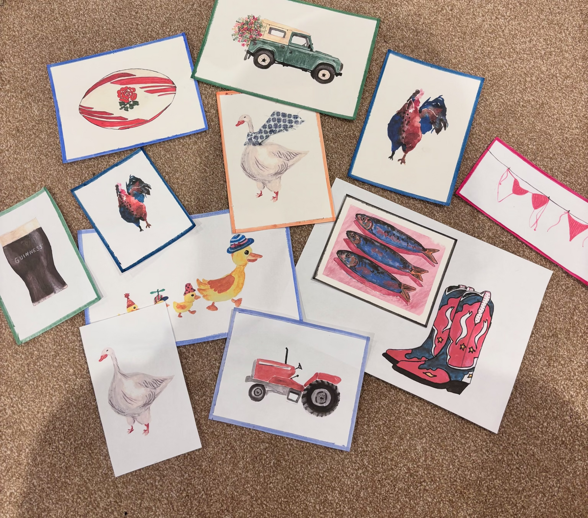 A collection of hand painted artwork arranged on a brown carpet. The artwork features whimsical drawings including a vintage Land Rover with flowers, a rooster, a goose wearing a scarf, a duck and ducklings wearing hats, mackerel, a tractor, cowboy boots, and a pint of Guiness.