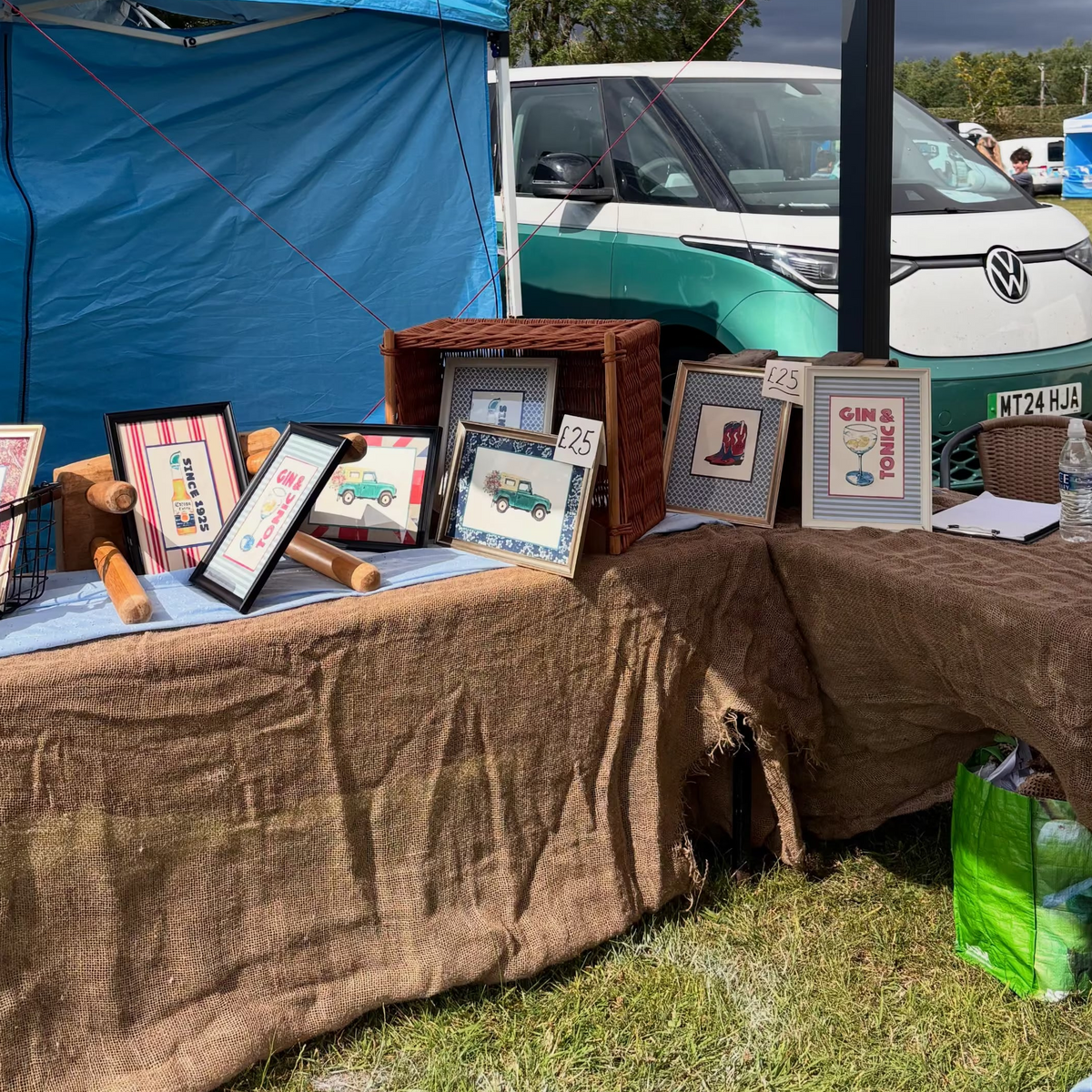 A market stall display featuring several framed art prints on a burlap-covered table, with a blue tent backdrop and a teal Volkswagen camper van in the background.